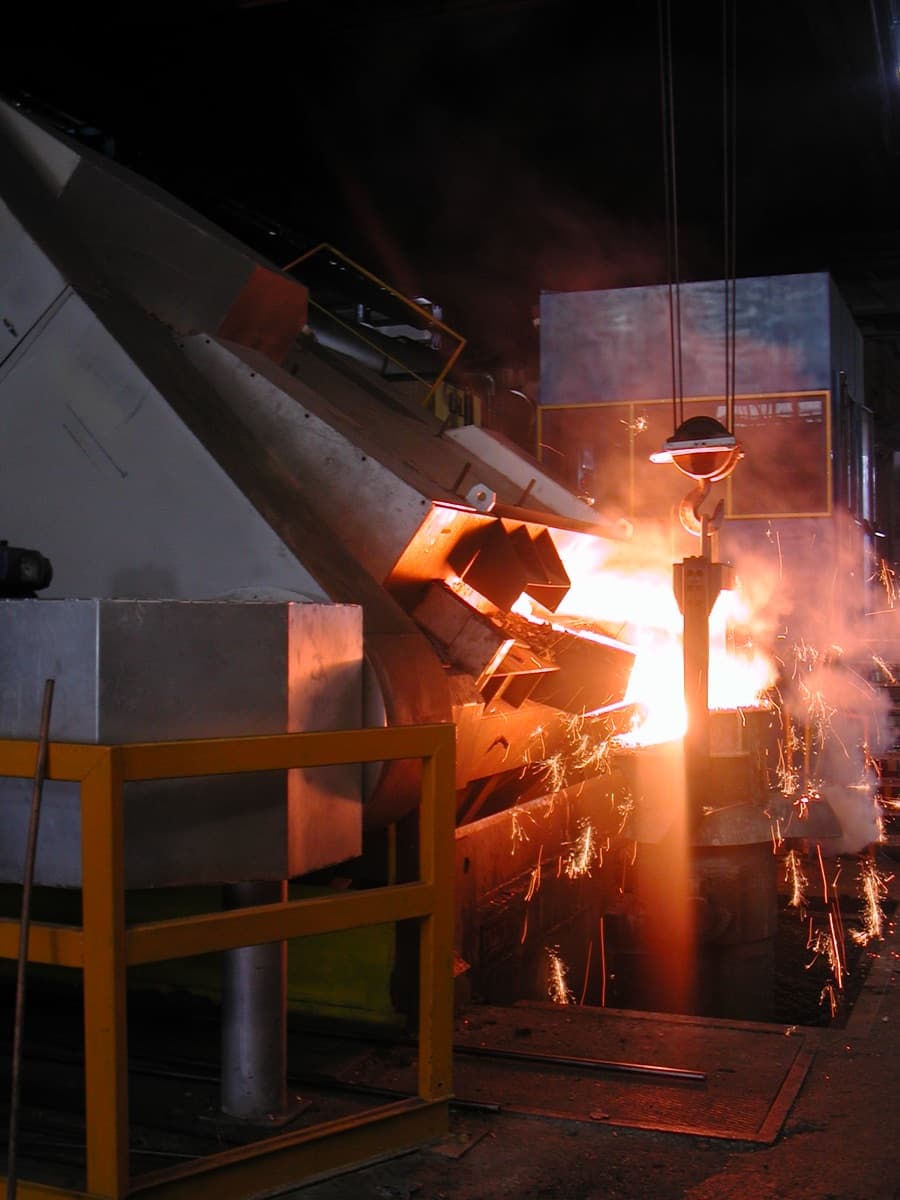 Tilted coreless induction furnace tapping molten metal, a bright stream of liquid iron and a shower of orange sparks in a darkened foundry