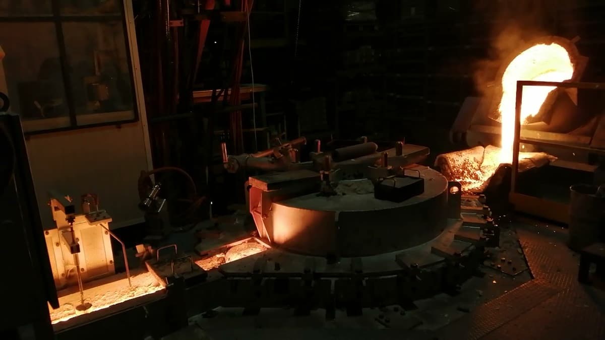 The sealed body of a CIME CAP press-pouring furnace clamped shut in a darkened foundry, a bright stream of molten iron charging the bath alongside